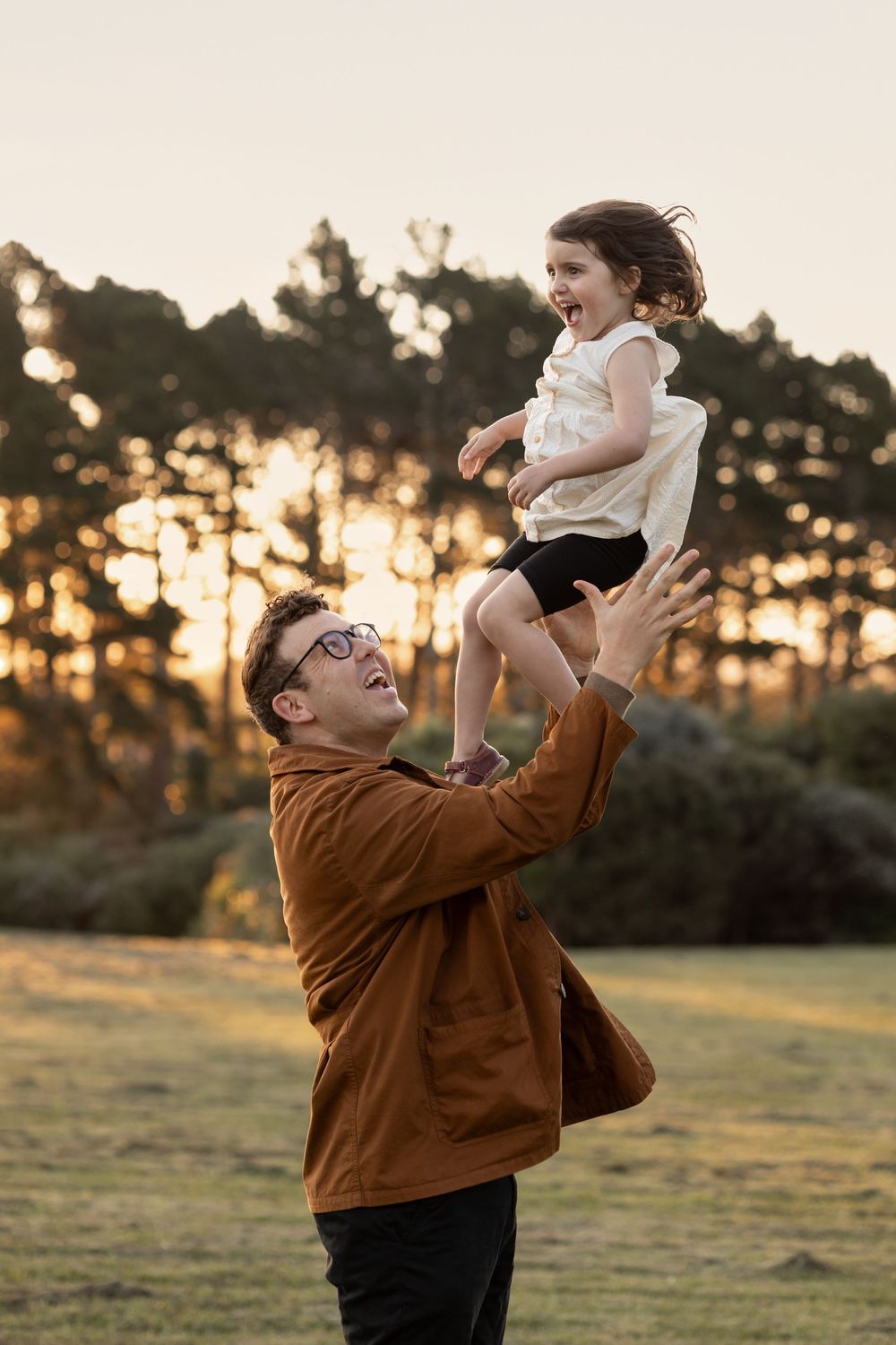 Father throws his young daughter playfully in the air for a family photoshoot at golden hour amongst the trees