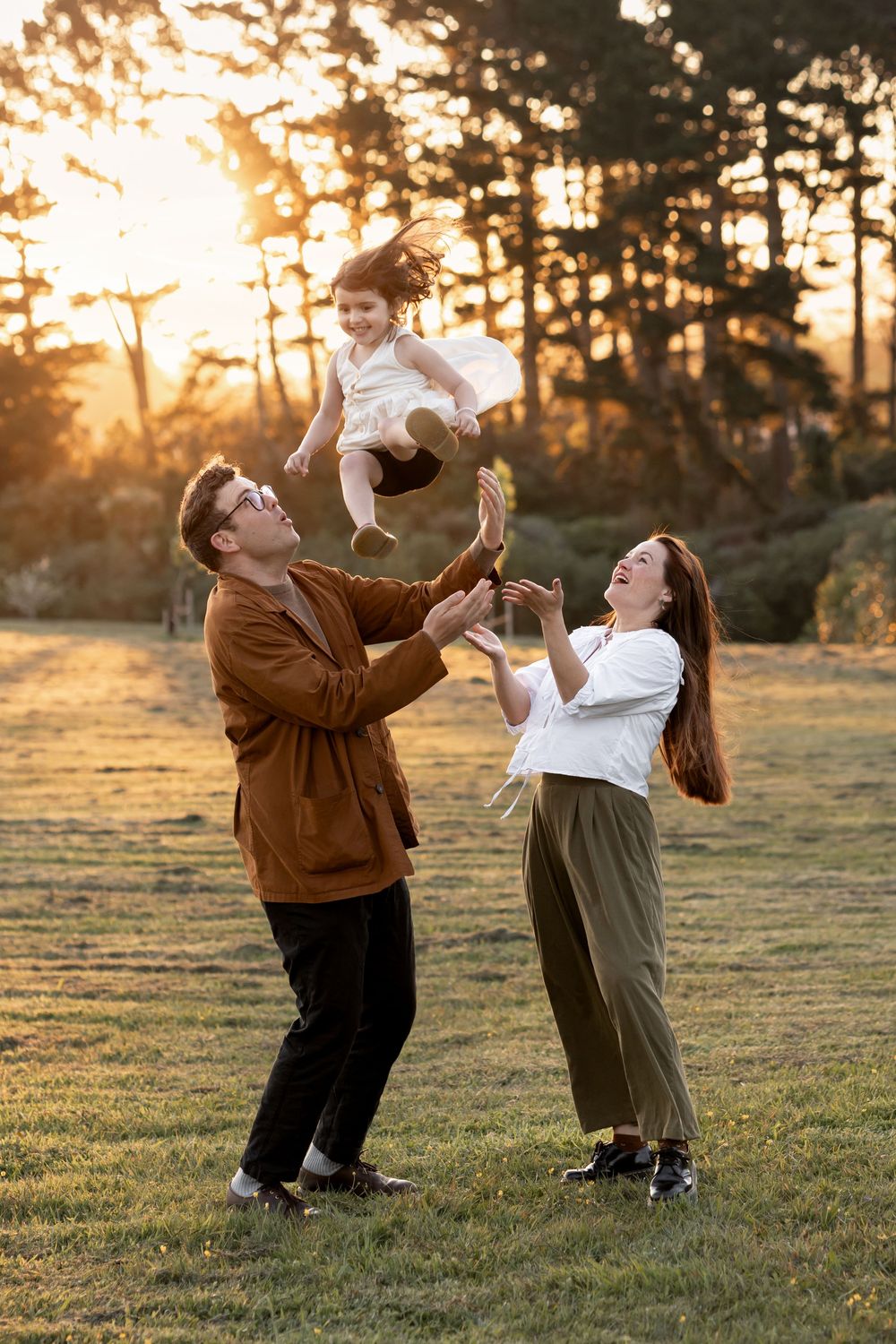 Family photoshoot in golden hour. Mother and father throwing daughter in the air for some fun family time. Sunset through the trees