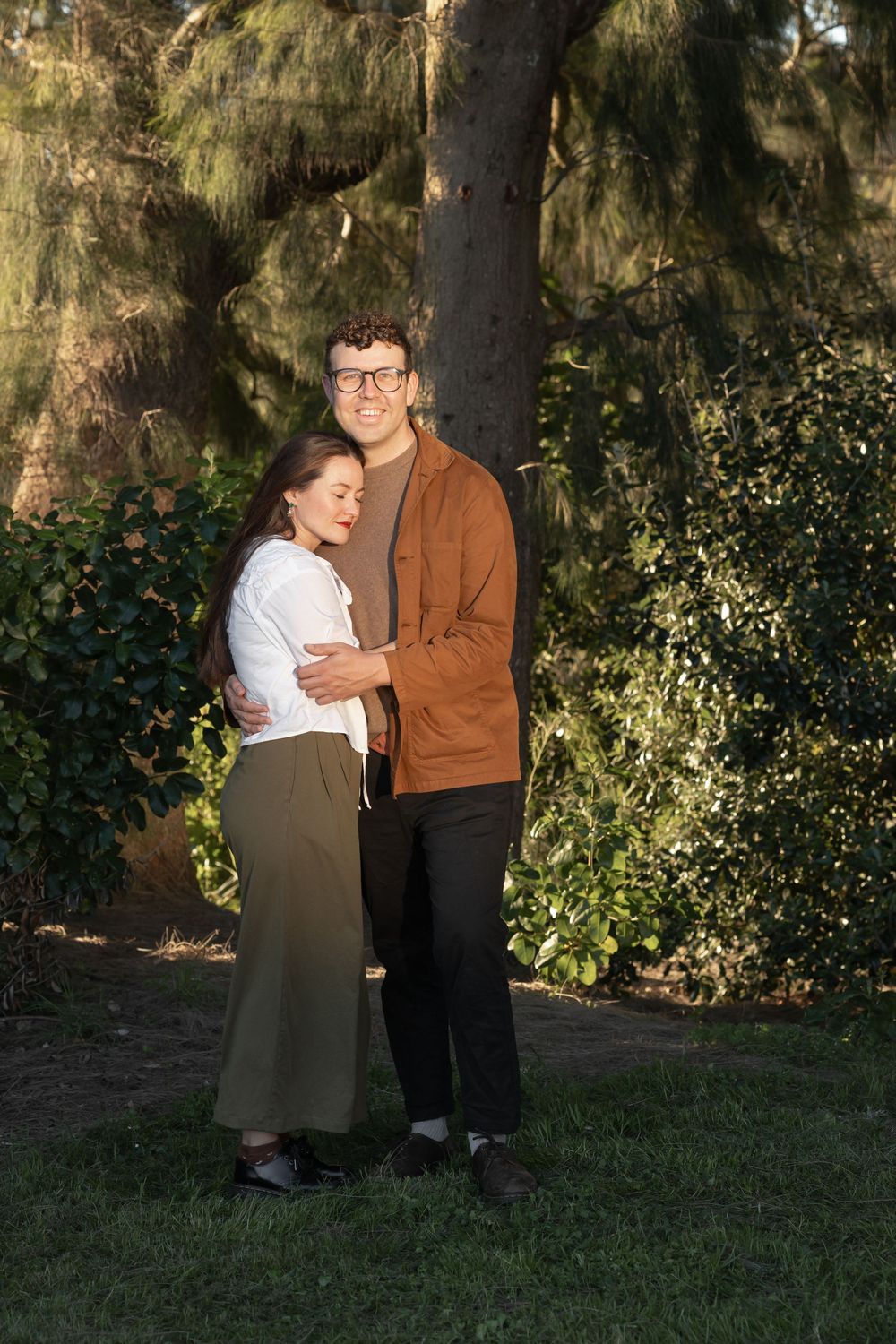 Mother and father embracing each other, standing in front of beautiful green trees with the sun shining on them at sunset