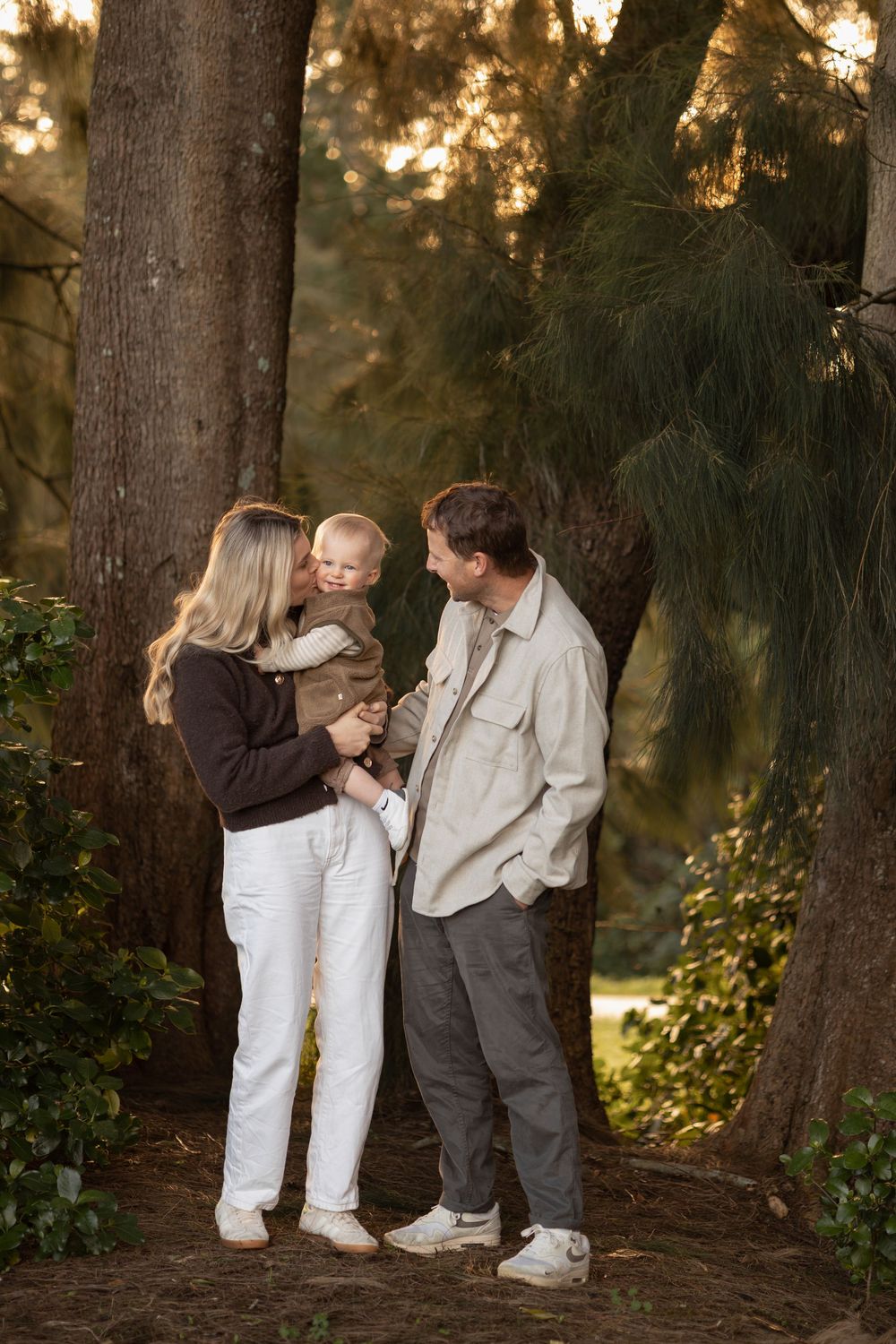 Golden hour family photoshoot in forrest with golden light shining through the trees, mother and father holds baby boy snuggling into him