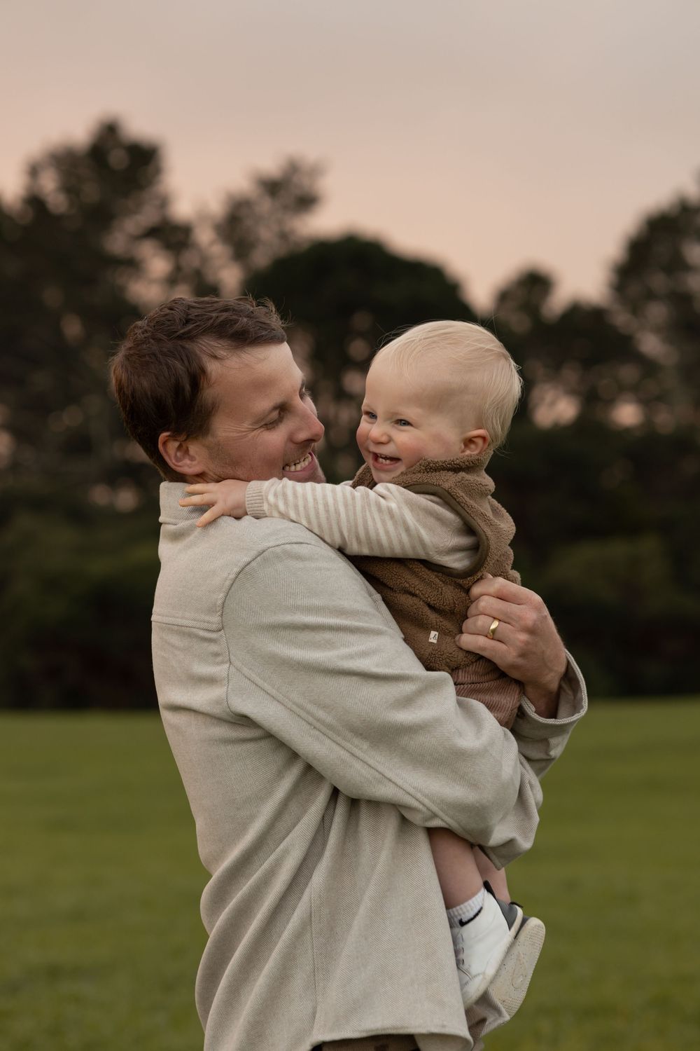 Golden hour with father holding his 1 year old boy they are laughing and snuggling together at sunset. Trees in background. Earthy tones