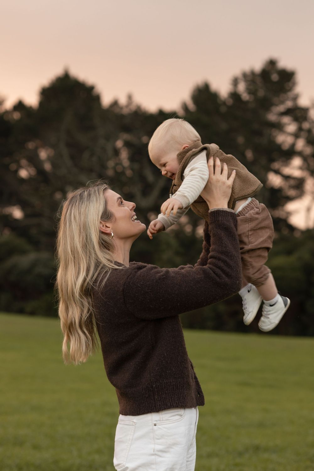 Mother holds her toddler up high with her blonde wavy hair flowing behind her. Golden hour family photoshoot . Greenery, trees. Laughters