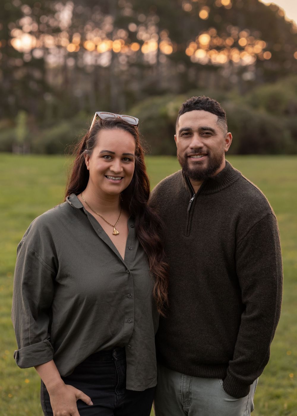 Professional Portrait of Husband and wife standing on grass field with large pine trees in the background. Sunset peeking through the trees