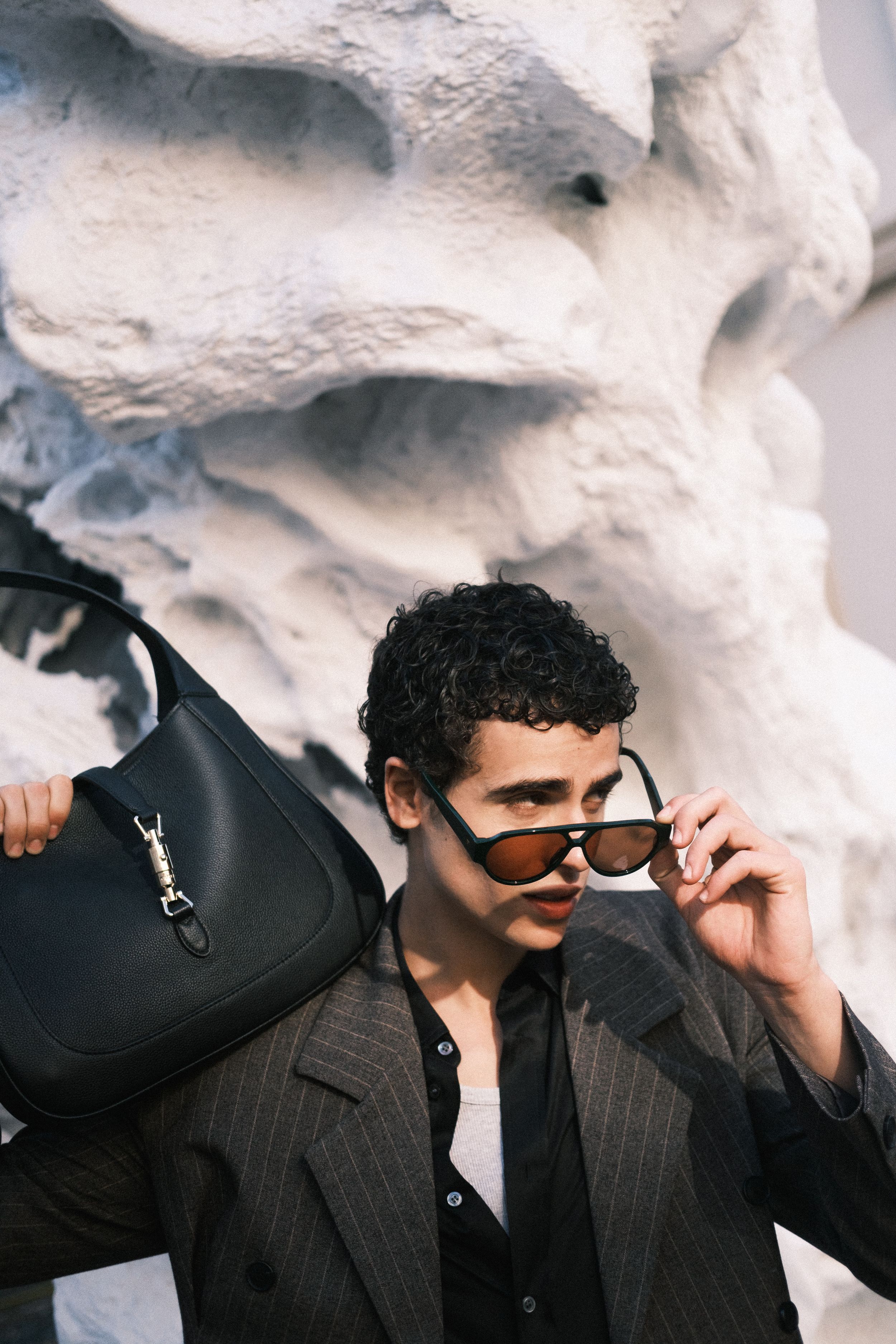 Fashionable person with curly hair adjusting sunglasses while holding a black leather bag.