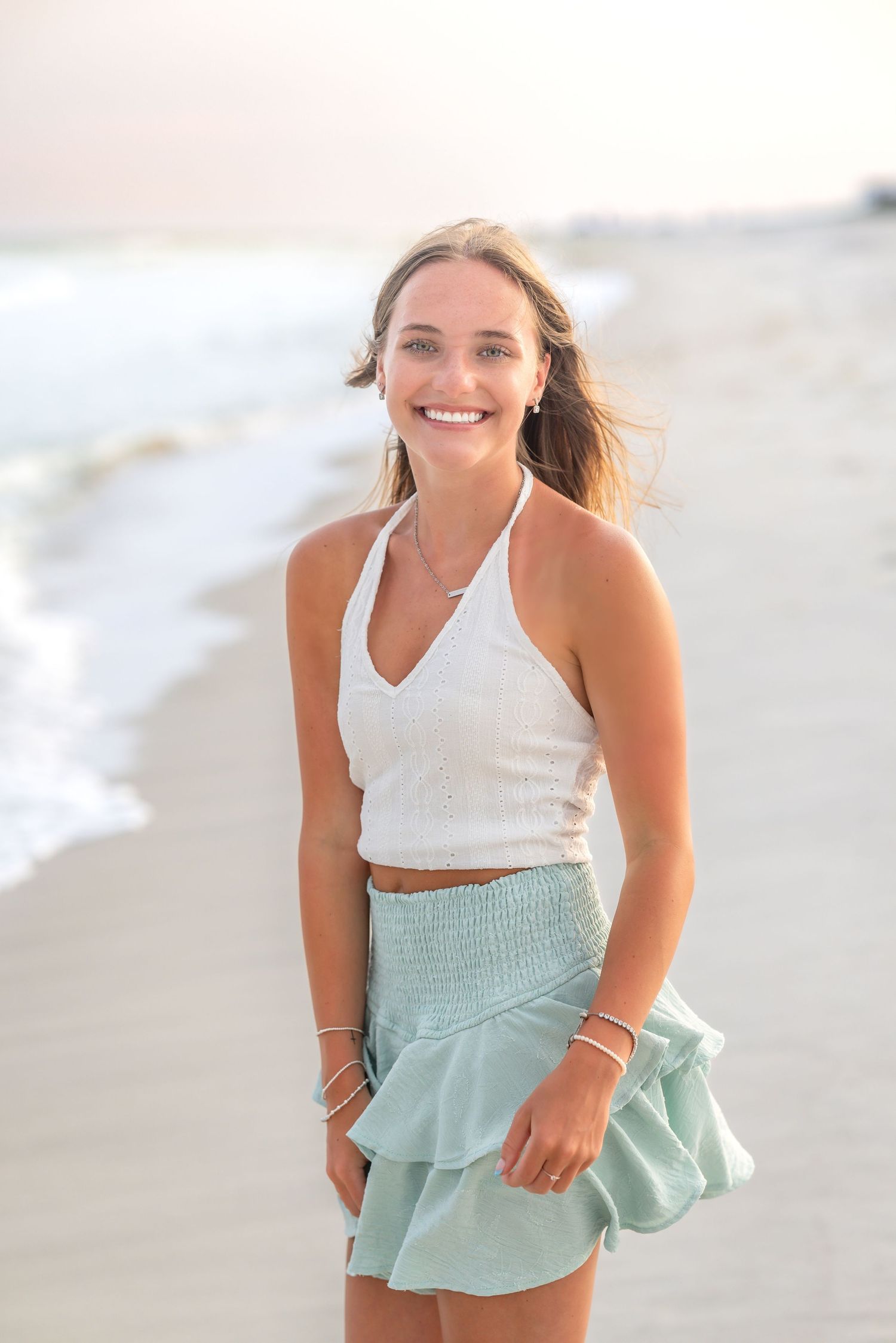 Someone wearing a white halter top and mint green ruffle skirt poses with a bright smile at the beach during sunset.
