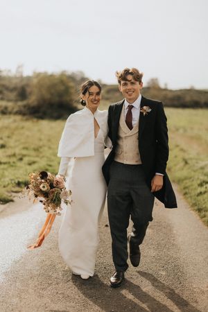 A couple in wedding attire strolls along a countryside dirt path near Askham Hall with warm sunlight creating a romantic atmosphere.