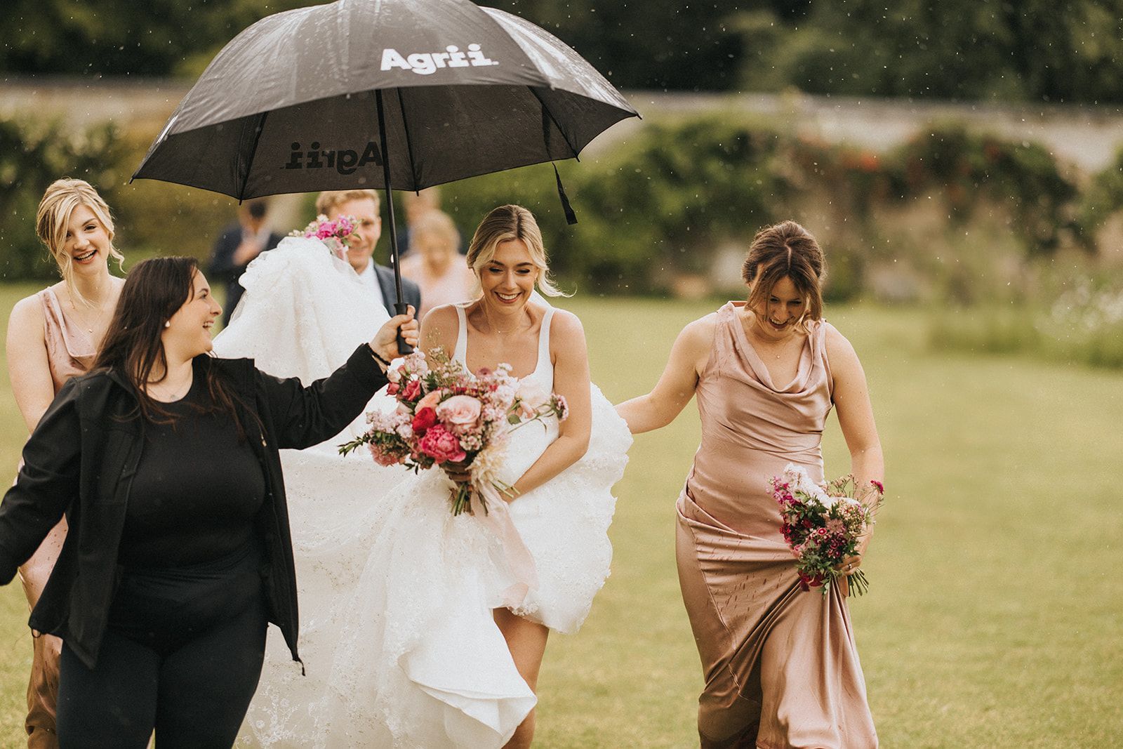 Wedding party members run through the rain holding umbrellas and bouquets on a grassy lawn.
