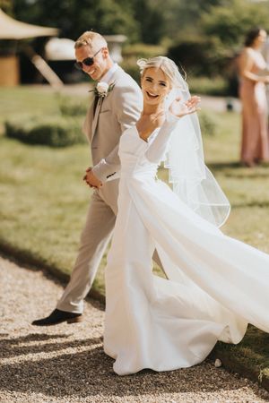A joyful wedding couple walks together outdoors on a sunny day at Hodsock Priory, with the bride in white and groom in beige suit.