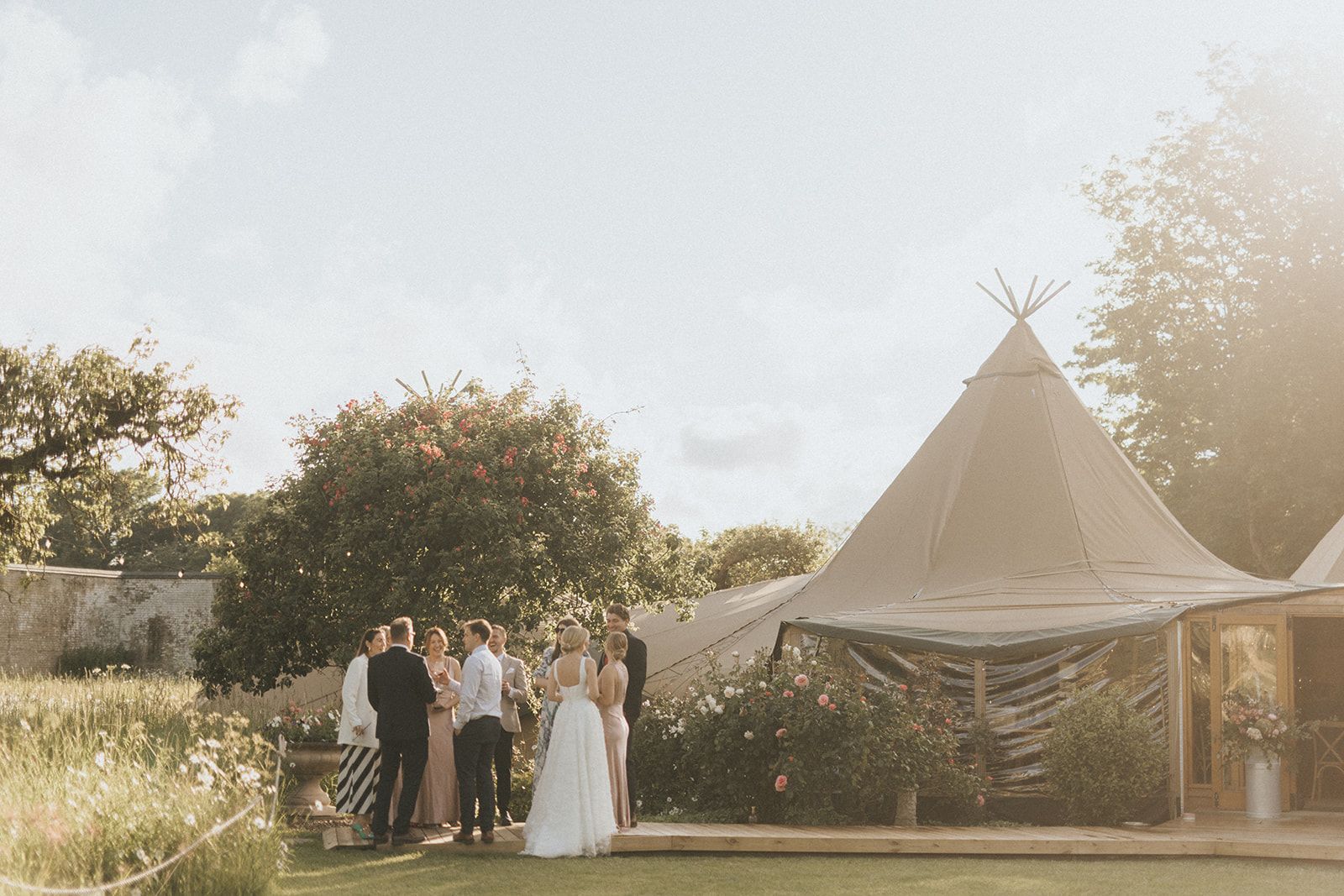Wedding party gathered outside a tipi tent structure in a sunlit garden setting during golden hour lighting.