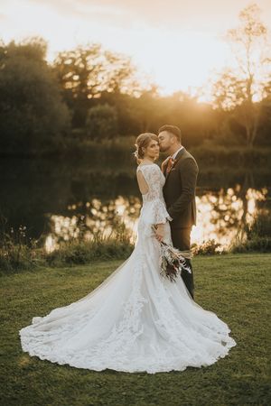 A romantic sunset wedding photo featuring a bride in a lace gown and groom embracing by a tranquil pond.