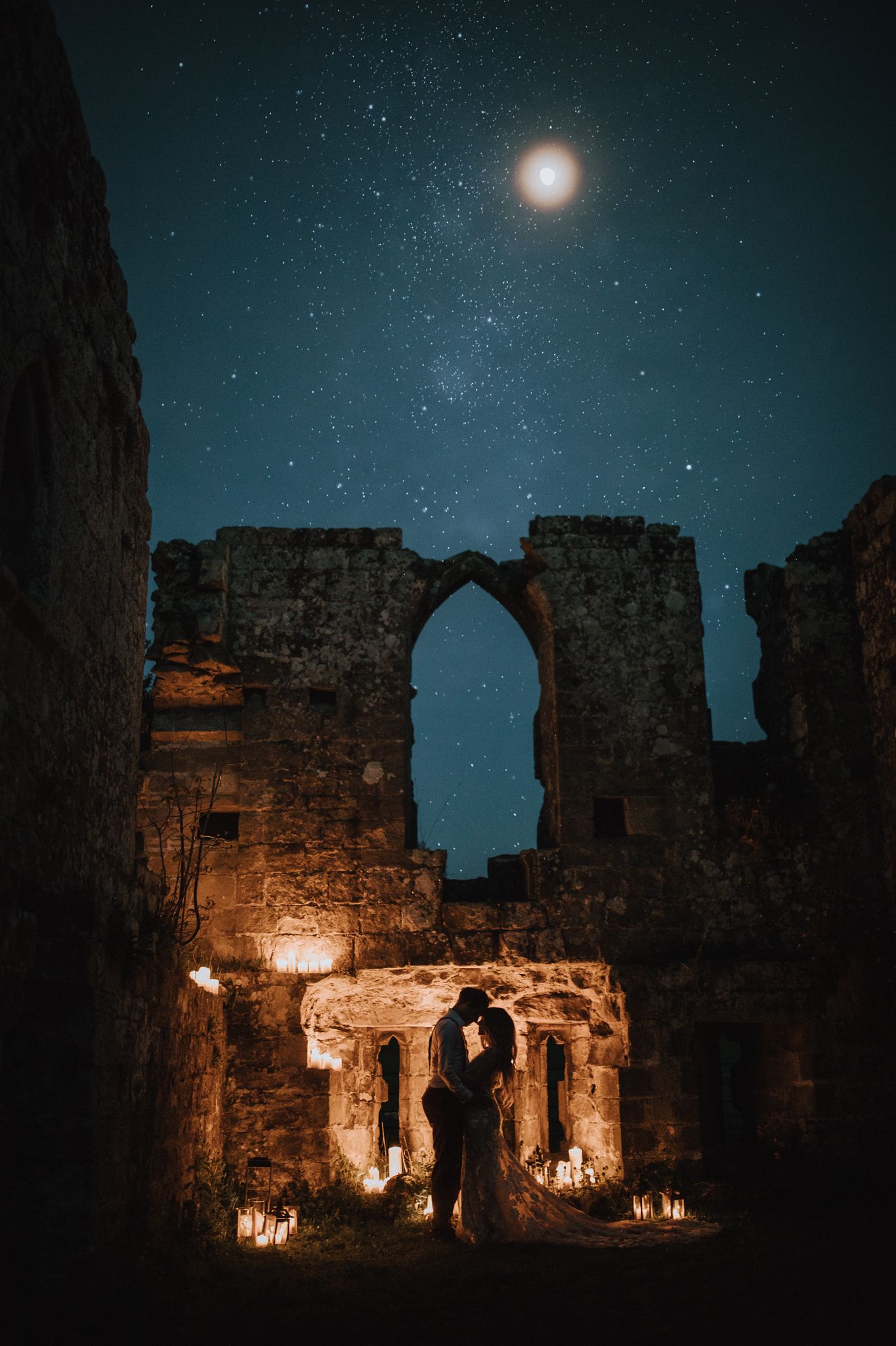 A couple embracing under a candlelit stone ruins and archway beneath a full moon and starry night sky creating a mystical atmosphere.
