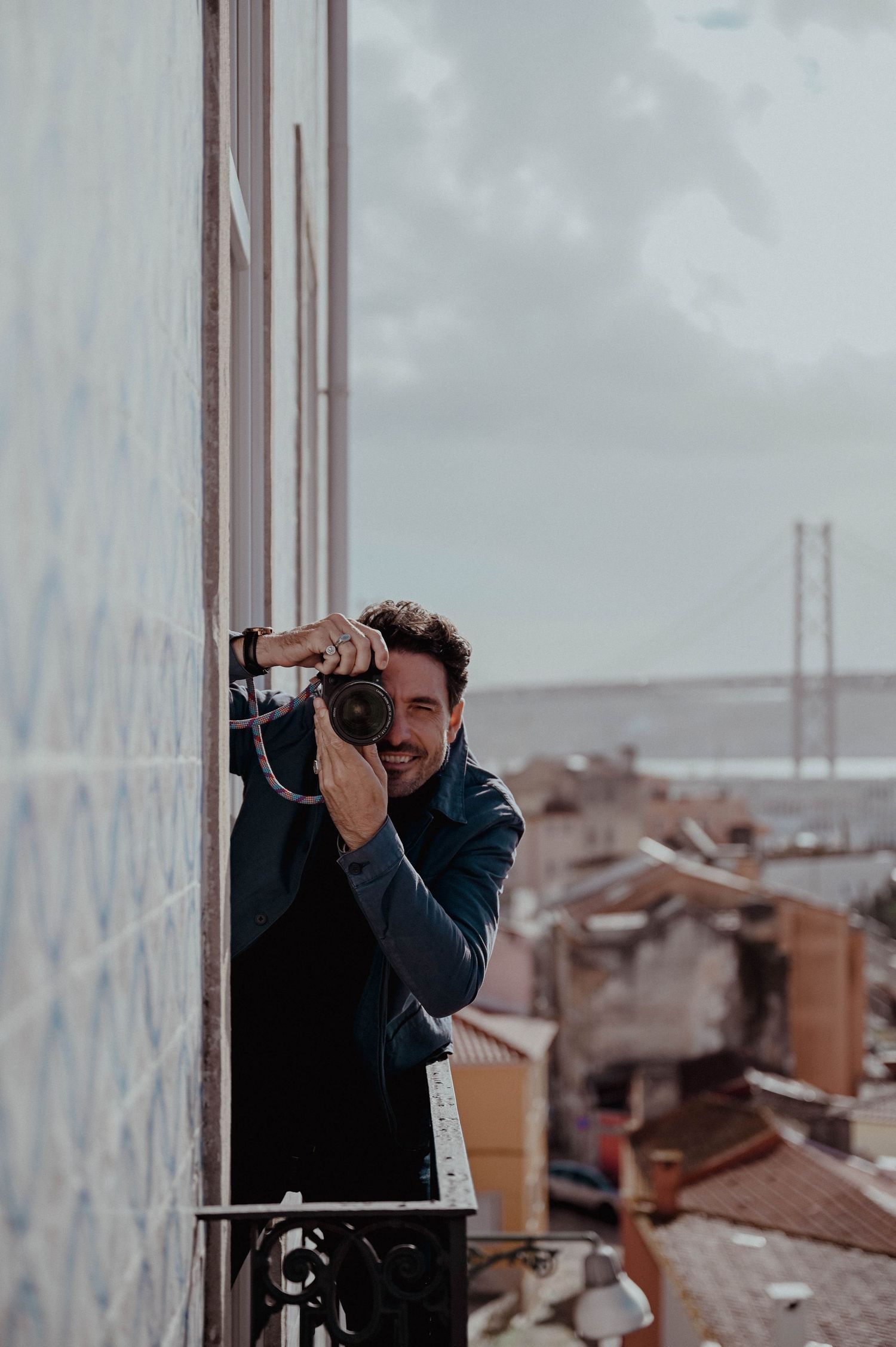 A wedding photographer in a dark coat takes photos from a high vantage point overlooking a cityscape with cloudy skies.
