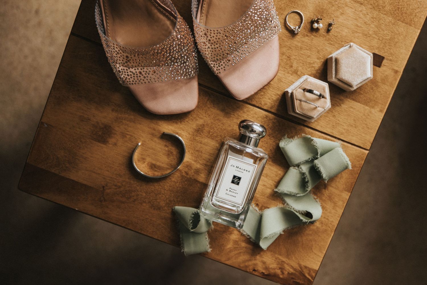 Wedding details flatlay with sparkly nude heels, perfume bottle, and sage green ribbons on wooden surface.
