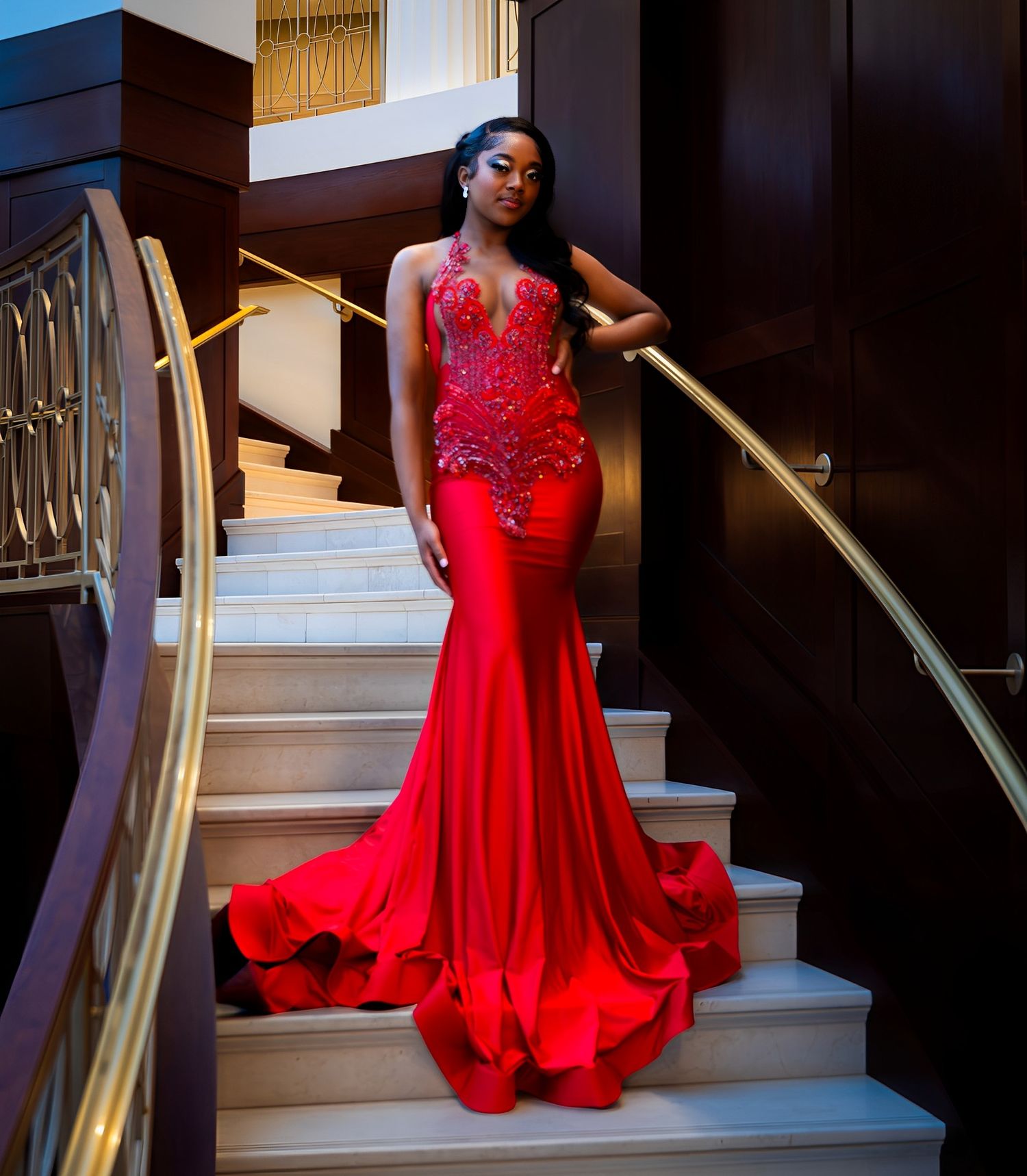A figure in a stunning red flowing evening gown poses elegantly on elegant stairs with gold railings.