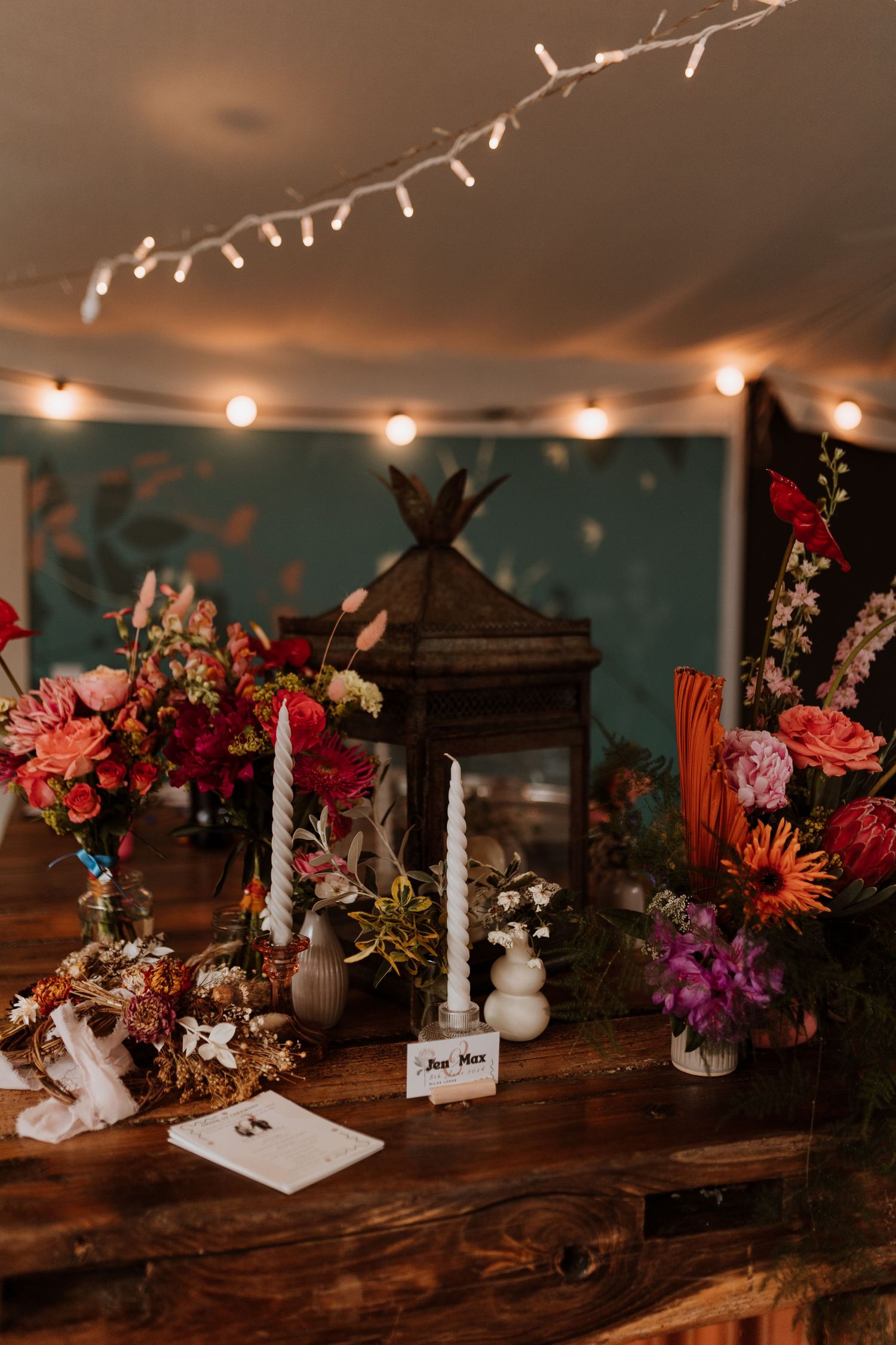 Wedding reception bar features rustic floral arrangements with pink and orange blooms on wooden surface with string lights above.