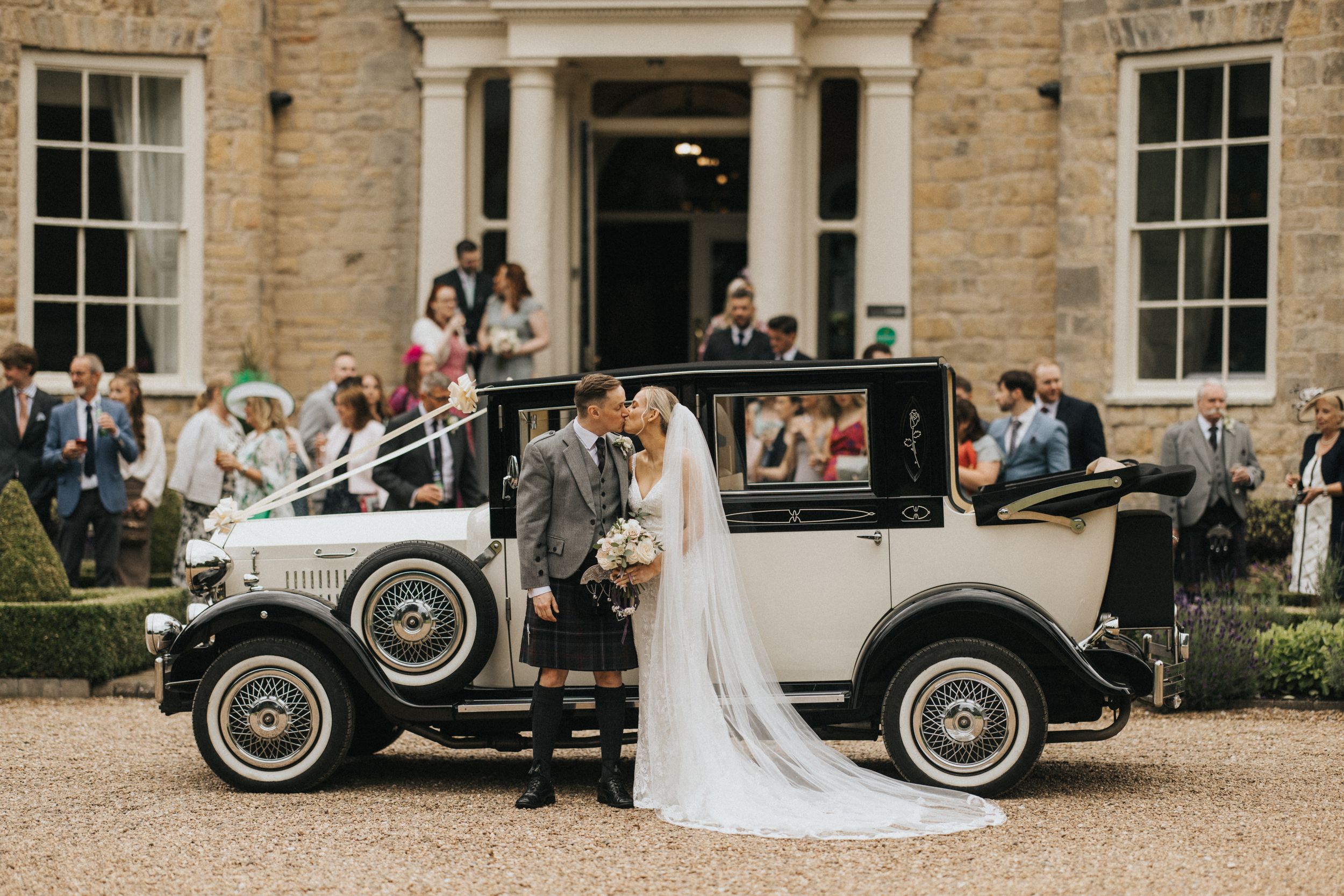 Newly weds kiss in front of vintage coloured classic car decorated for a wedding outside Washingborough Hall - with guests gathered around.