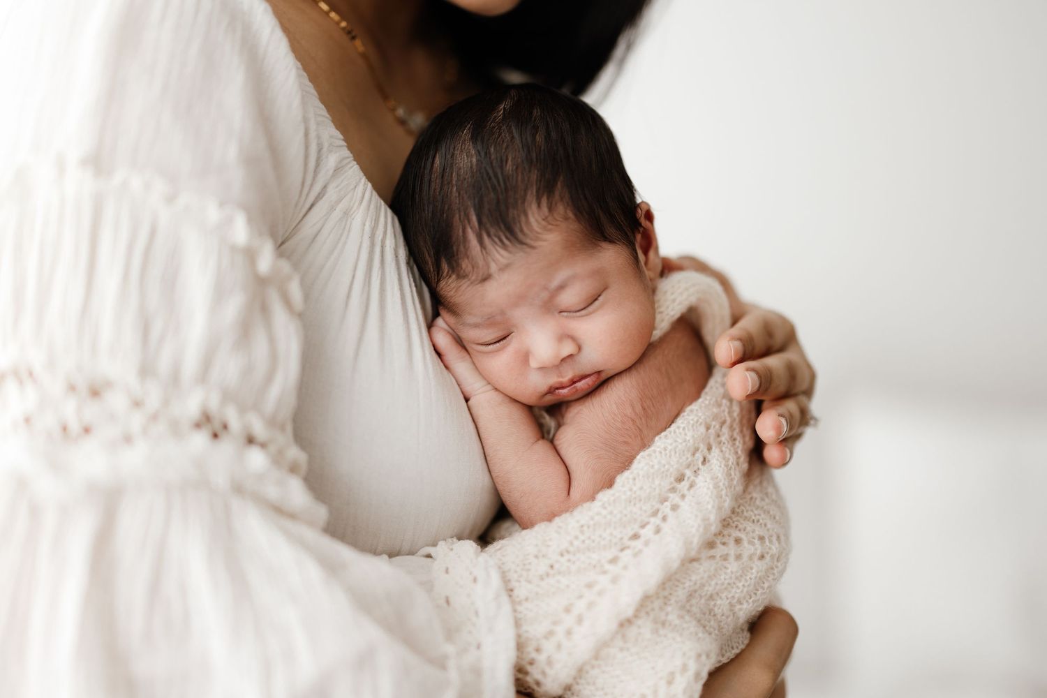 A newborn baby wrapped in a white knit blanket sleeps peacefully against a figure in a white lace dress.