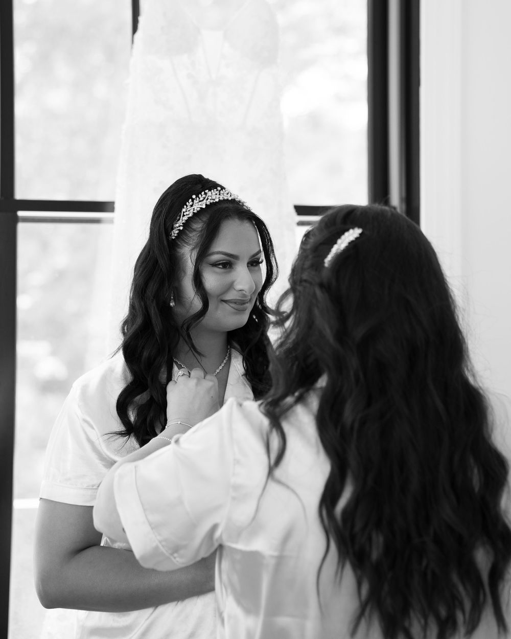 Two women in white shirts are reflected in mirrors while doing hair and makeup styling.
