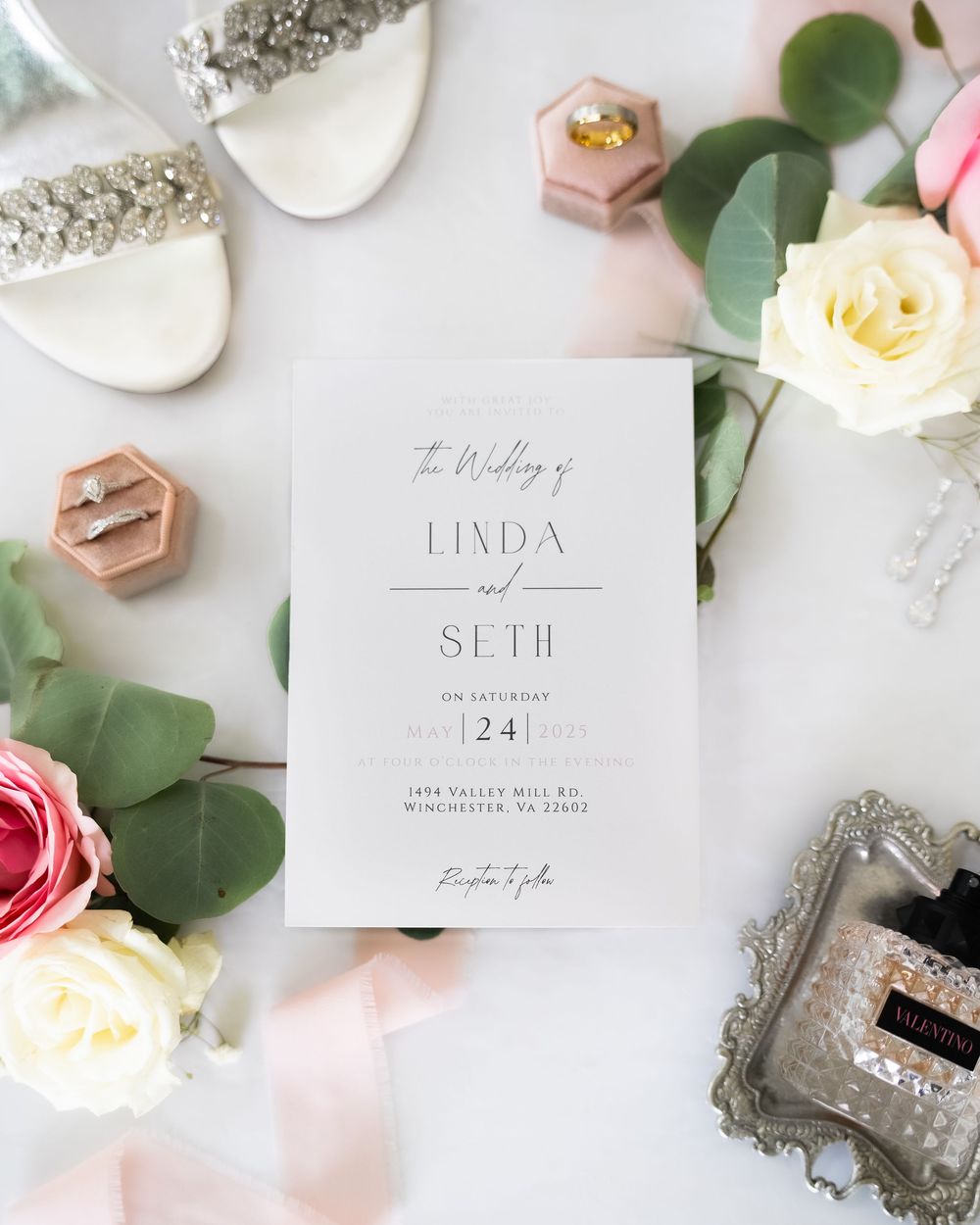 Elegant wedding invitation with white floral accents surrounded by roses, eucalyptus leaves and bridal accessories.