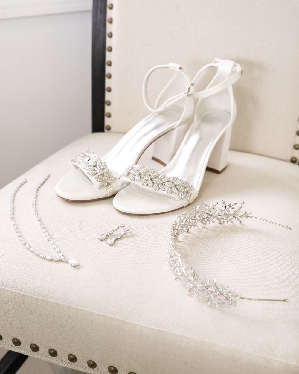 Elegant white bridal shoes displayed alongside sparkling crystal jewelry pieces on a white cushioned surface.