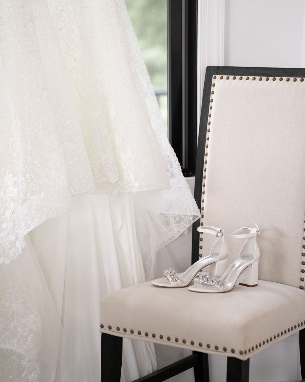 Delicate white lace and tulle fabric details of a wedding dress hanging from black chairs against a light background.