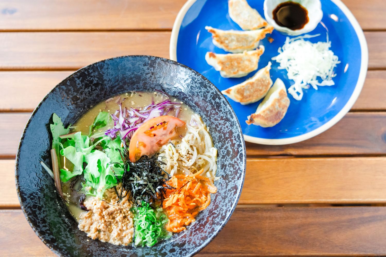 Multiple bowls of colorful Asian rice and noodle dishes with vegetables arranged on a wooden table with blue plates.