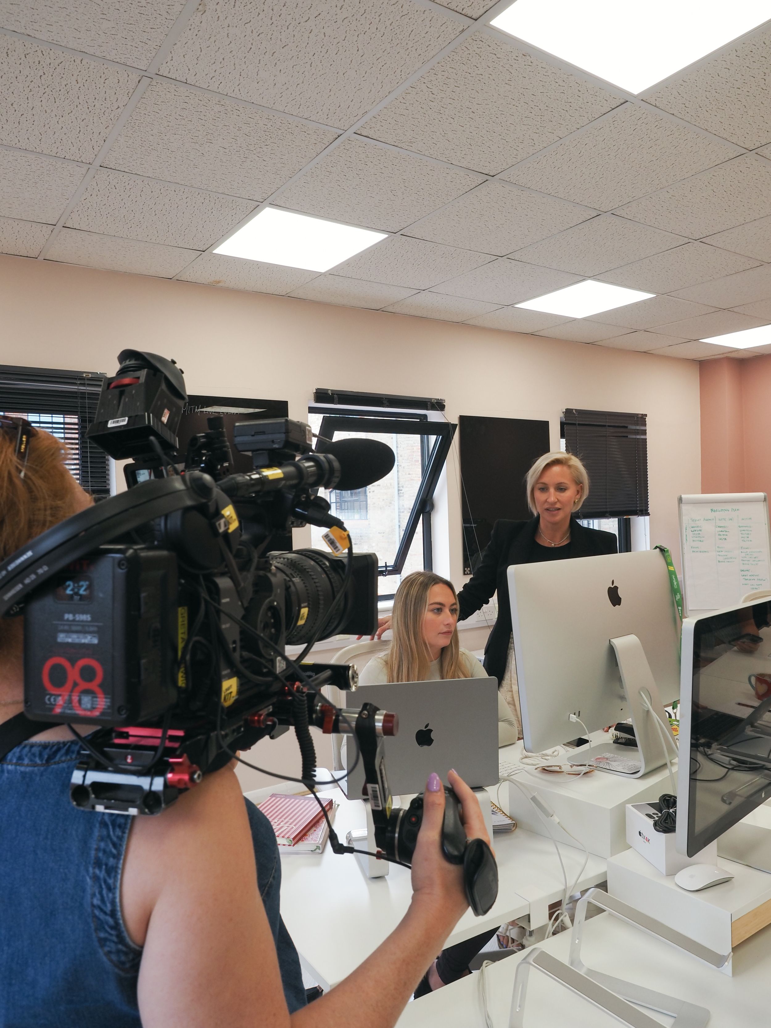Camera operators film content at workstations in a bright office setting with multiple computer displays.