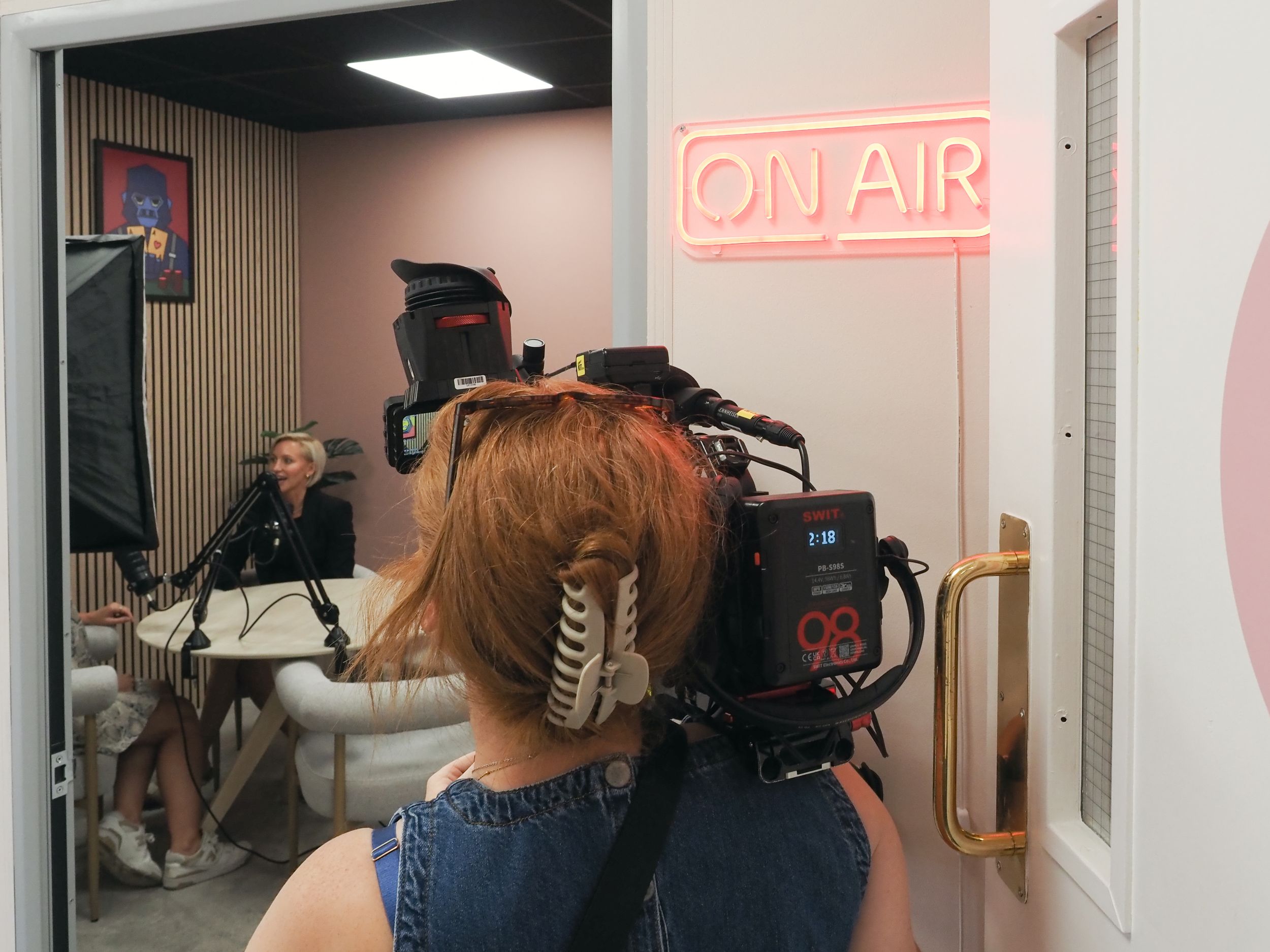 A camera operator films in a small recording studio with an illuminated 'ON AIR' sign visible in the background.