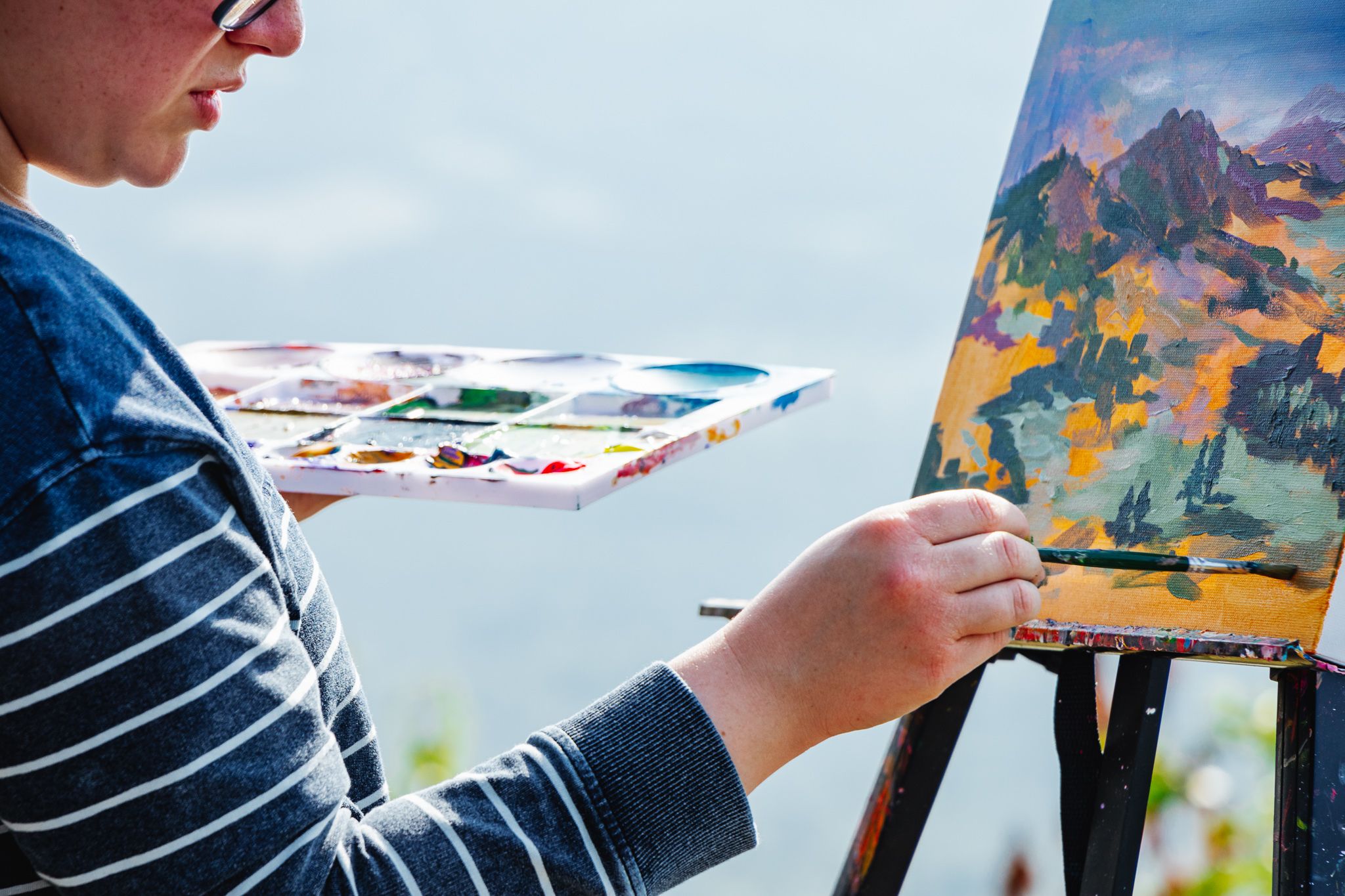 A close-up of an artist in a striped shirt concentrating on painting a landscape, holding a paint palette and working on canvas outdoors.