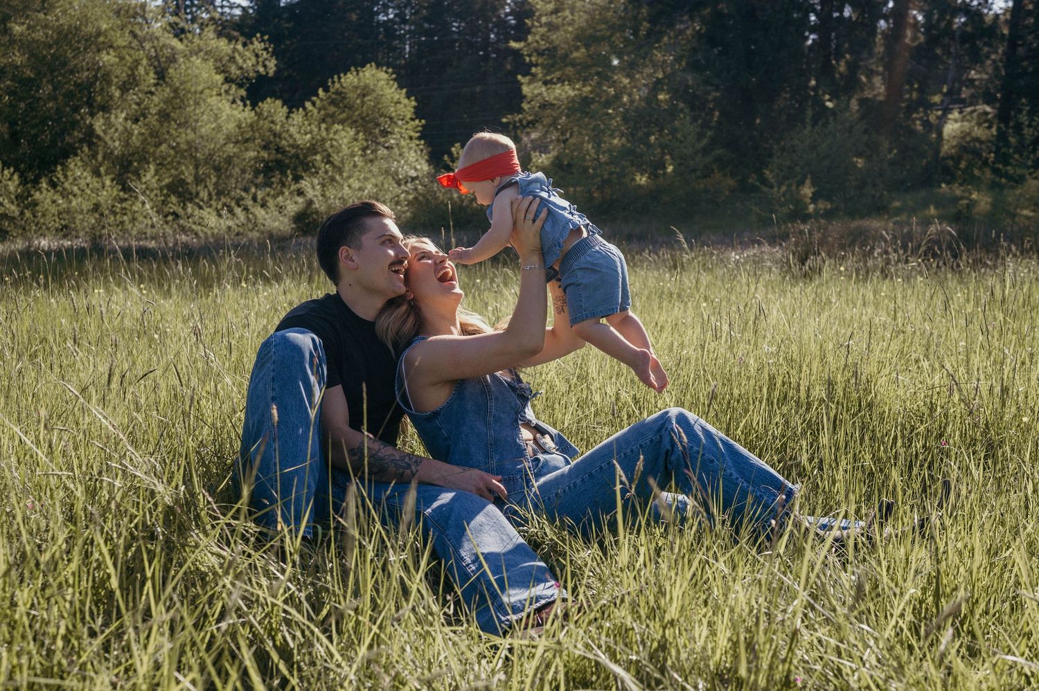 A happy young couple sits in a lush green meadow in Port Orchard Washington, playfully lifting their baby in the air on a sunny day.