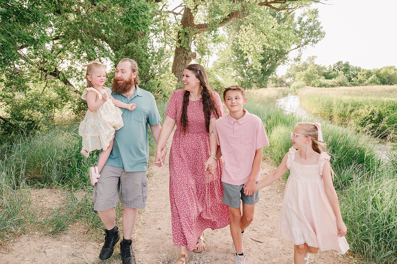 A family walks together down a tree-lined dirt path on a sunny day in nature.