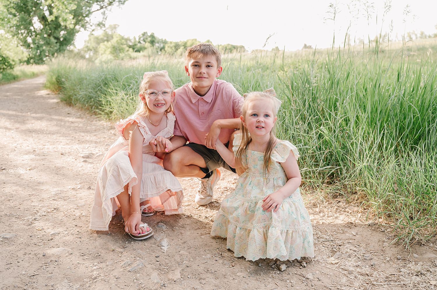 Three children in light colored clothing crouch together on a dirt path surrounded by tall summer grasses.
