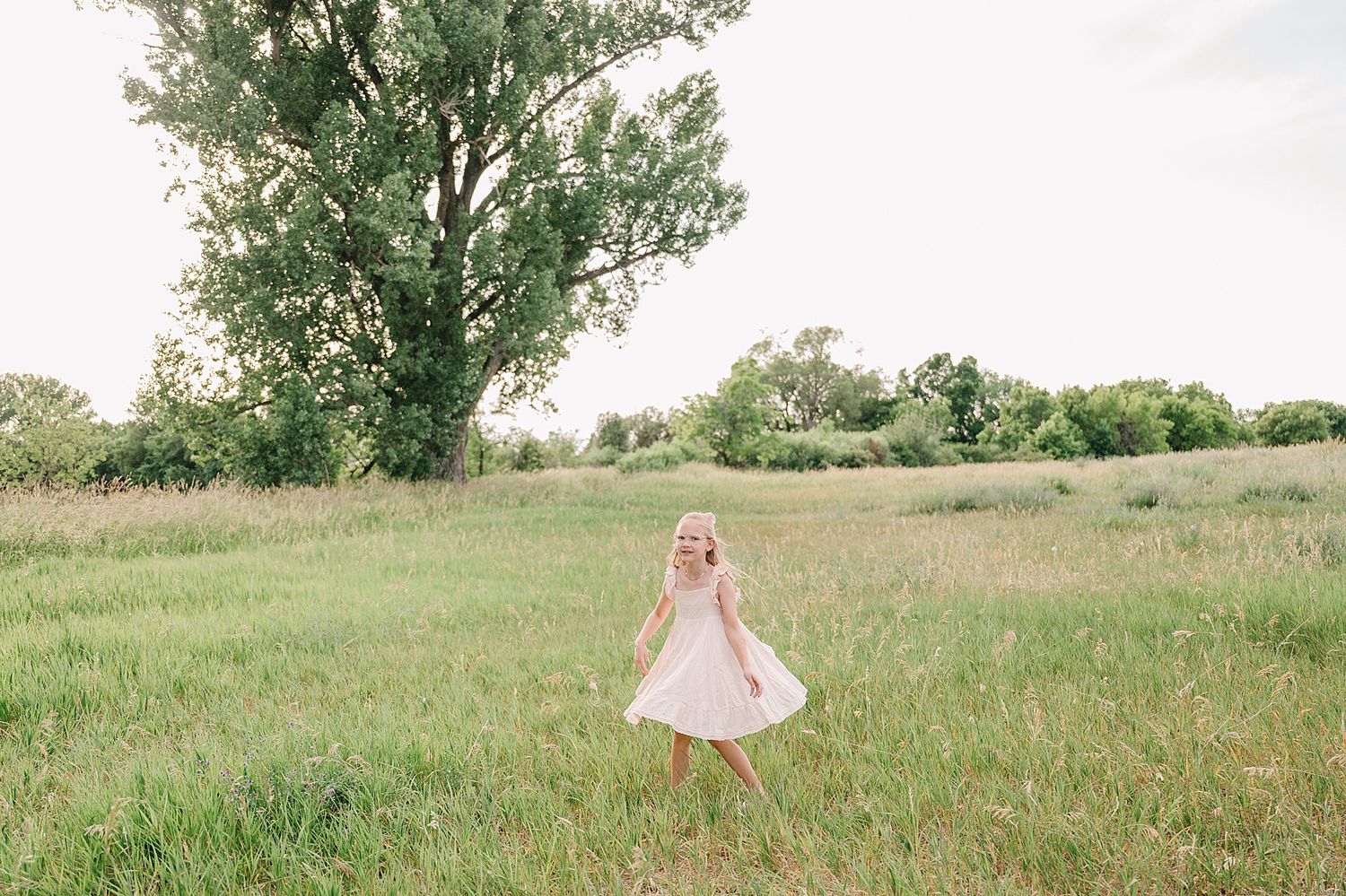 A person in a flowing cream dress twirls in a sunlit meadow with tall grass and trees in the background.