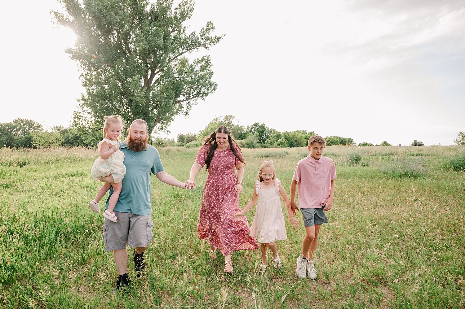 Candid lifestyle family portrait session in a sunlit meadow with parents and children walking together through tall grass.