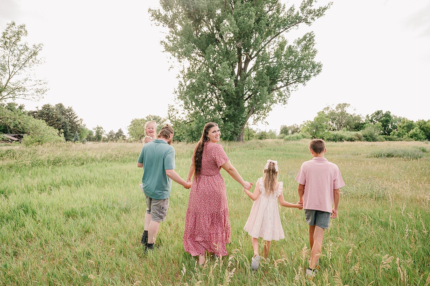 Family walking together through a lush green meadow with tall trees in the background on a bright summer day.