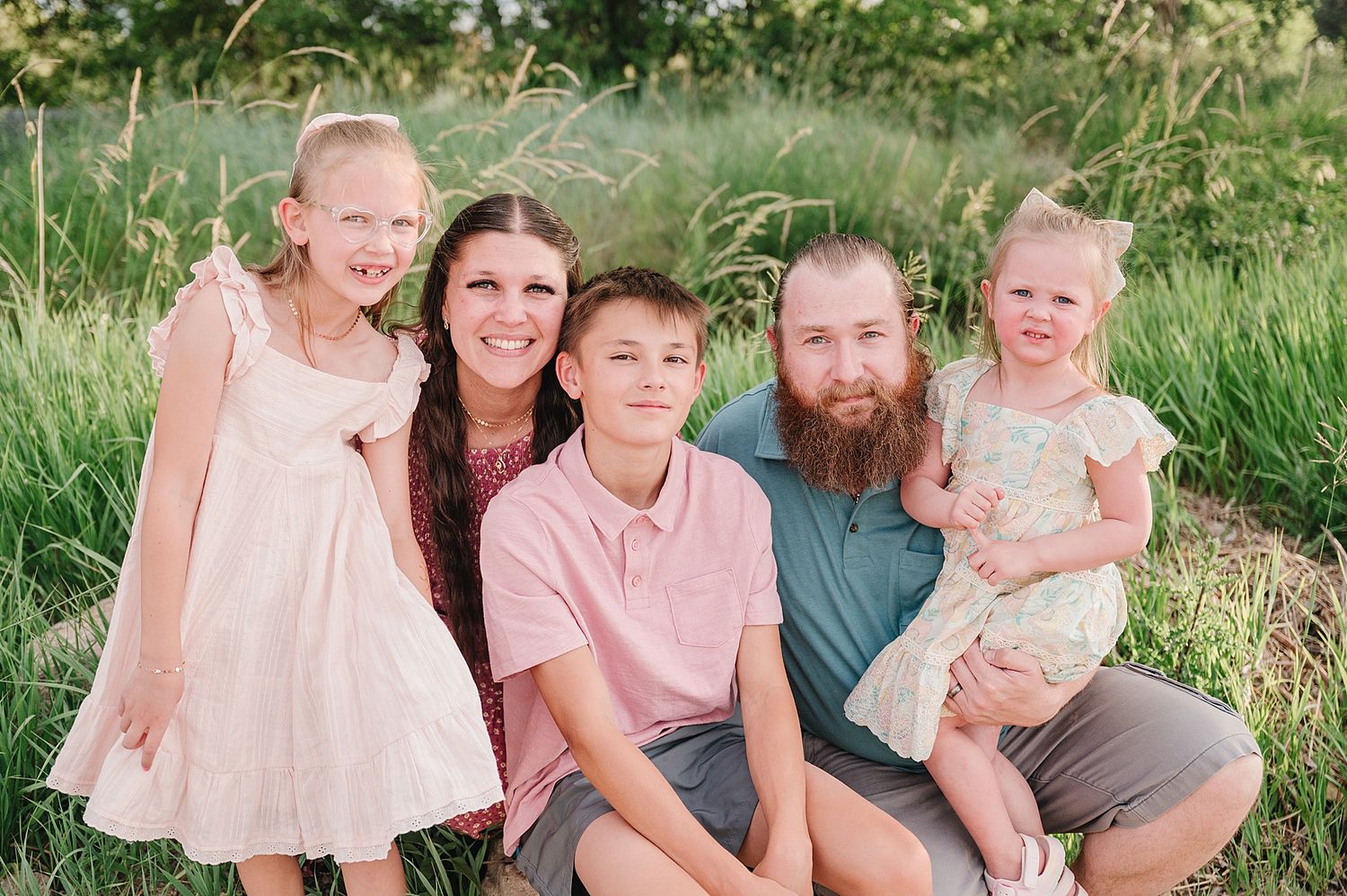 A family in coordinated pink and grey outfits sits together on grass during an outdoor portrait session.