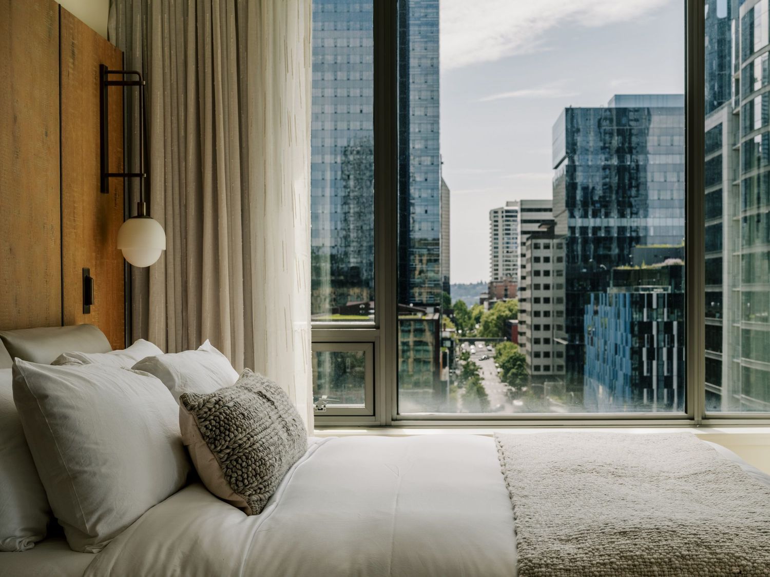 Modern bedroom with floor-to-ceiling windows offering panoramic city views, white bedding and a plush decorative pillow.