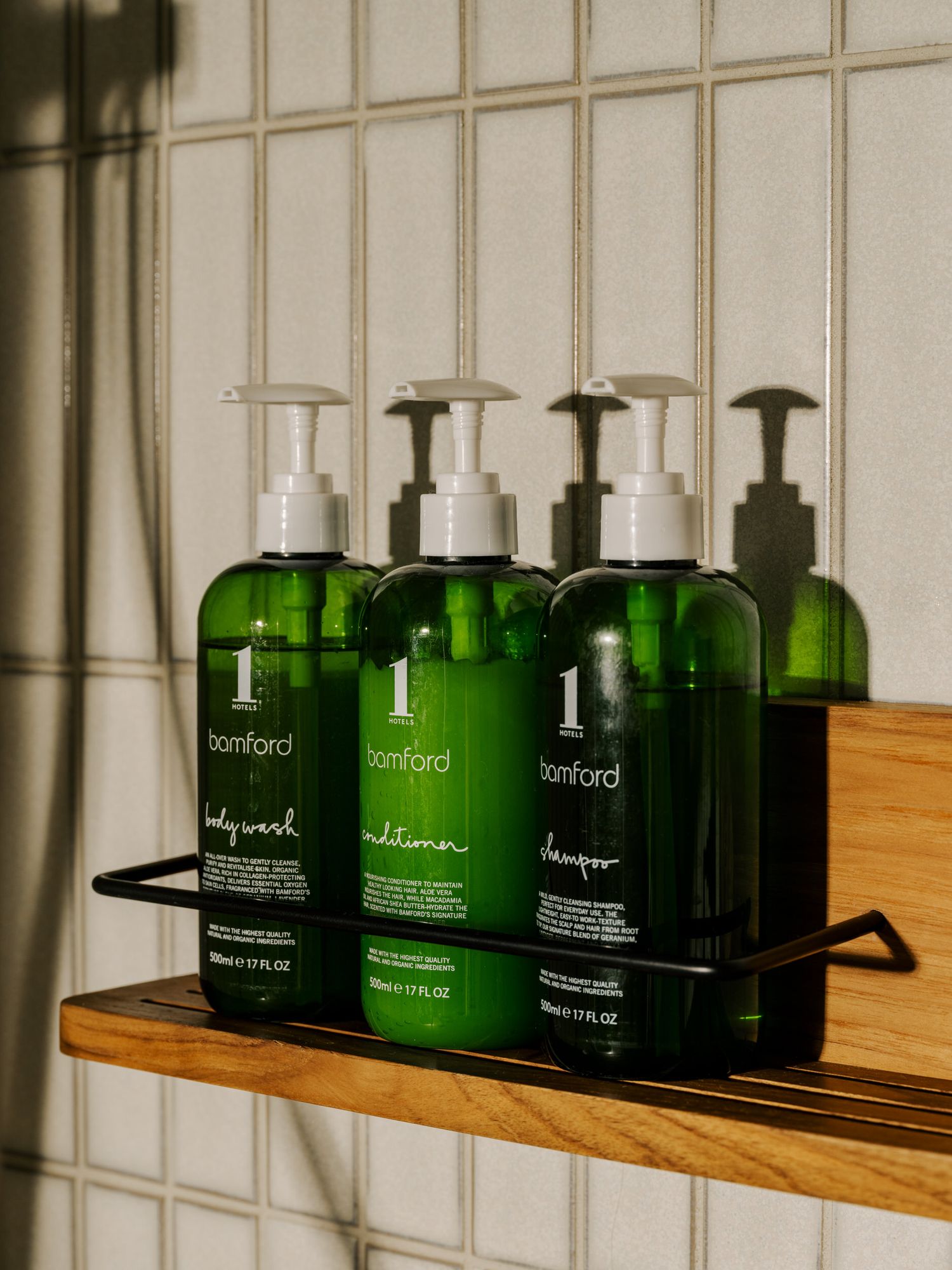 Green glass bottles of luxury bath products arranged on wooden floating shelves against a white tiled wall.