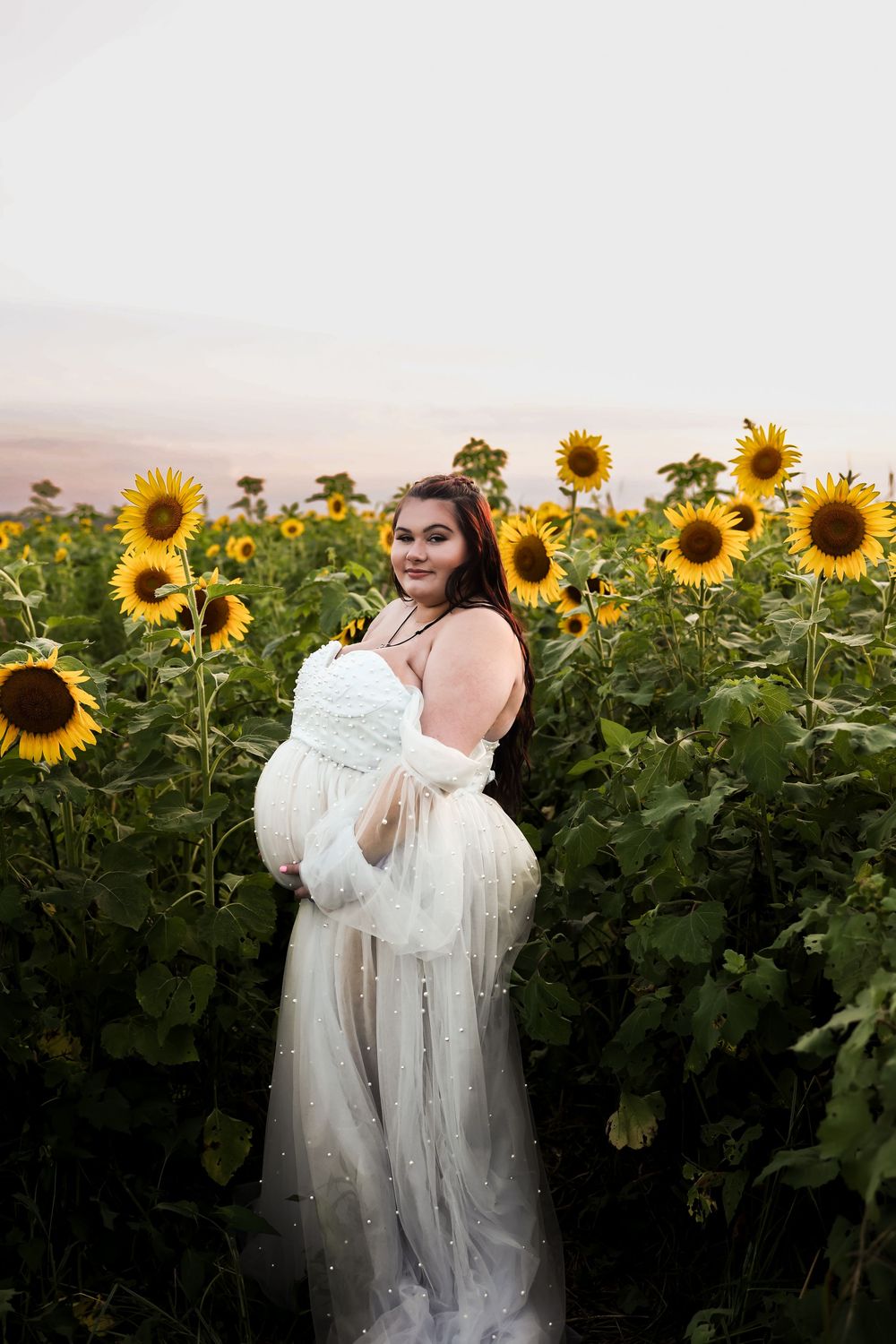 Person in a flowing white dress poses in a blooming sunflower field at sunset with glowing golden light.