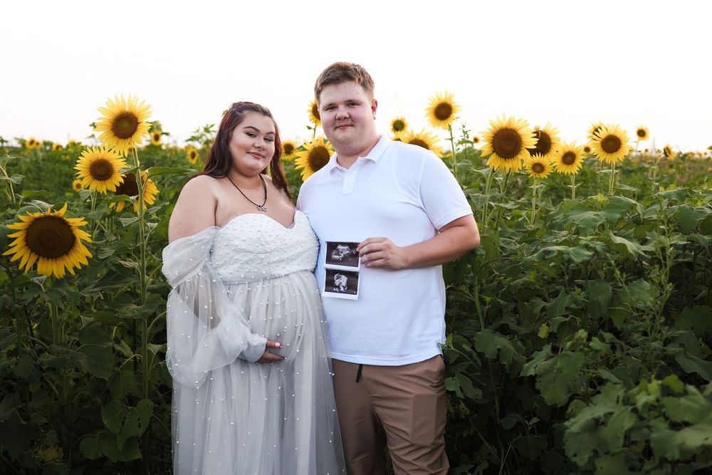 A couple holding ultrasound photos poses together in a beautiful sunflower field at sunset.