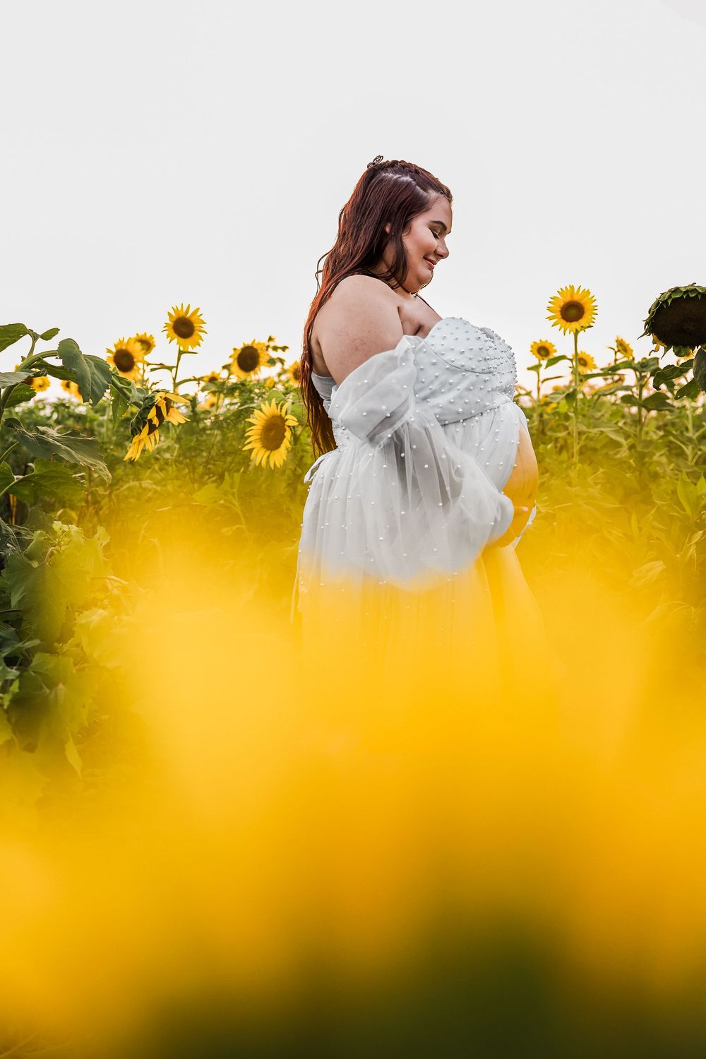 A pregnant woman in a white dress stands among blooming sunflowers in a golden sunlit field.