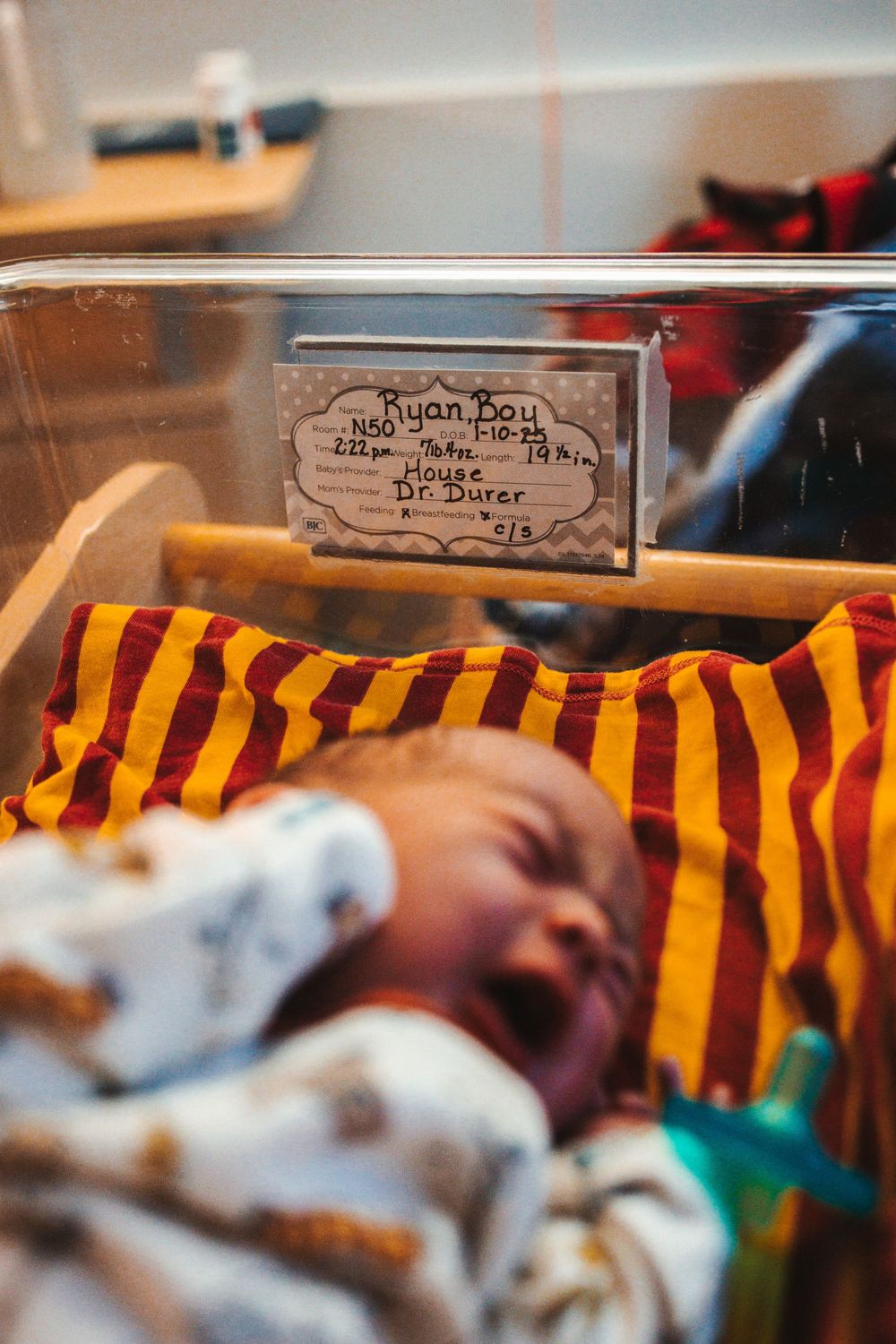 Hospital card and red and yellow striped blanket visible in newborn nursery setting through glass window.