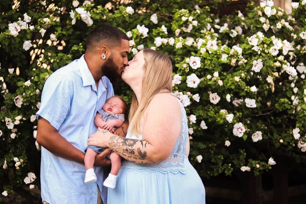 A couple in blue and white outfits poses for romantic engagement photos among blooming white flowers in a garden setting.
