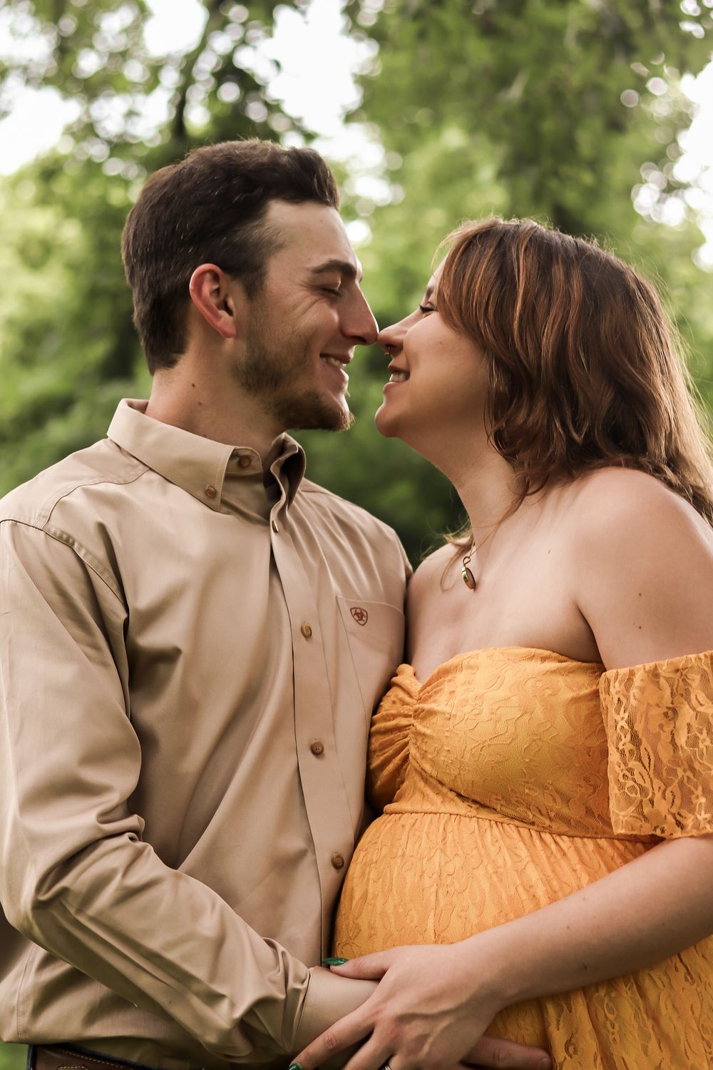 A romantic couple shares a tender moment outdoors as they celebrate their upcoming parenthood in matching yellow and tan outfits.