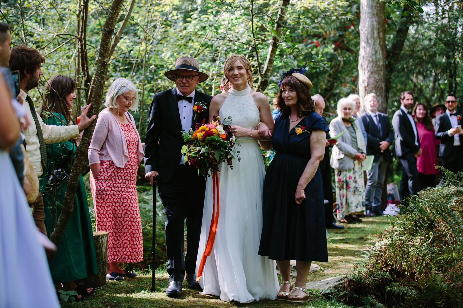 A wedding ceremony takes place on a wooded path with guests gathered among lush greenery.