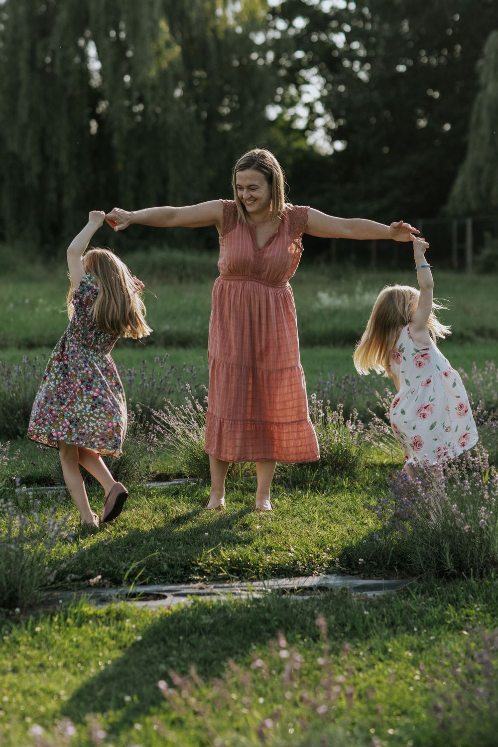 Mother playing with two daughters in floral dresses among blooming garden flowers.