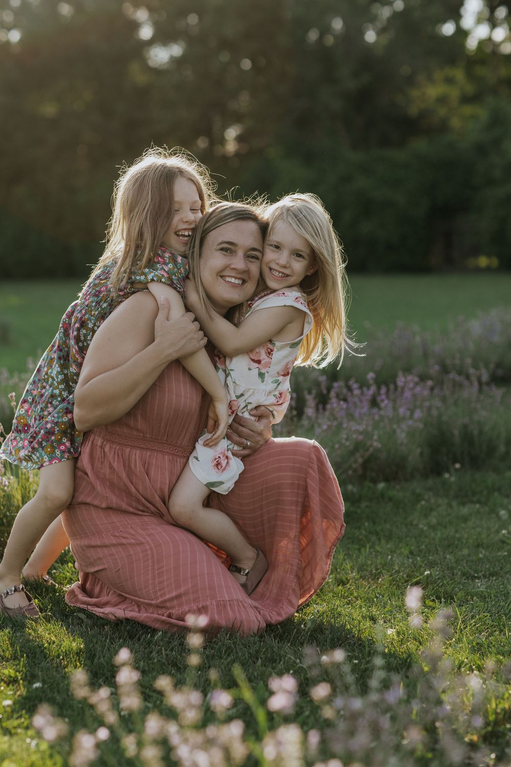 Mother and daughters share loving embrace while kneeling in garden at sunset.