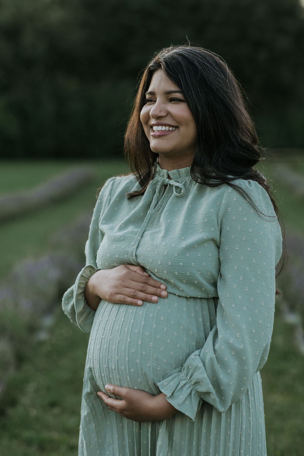 Pregnant woman in mint green dress smiles while cradling her baby bump in a garden setting.
