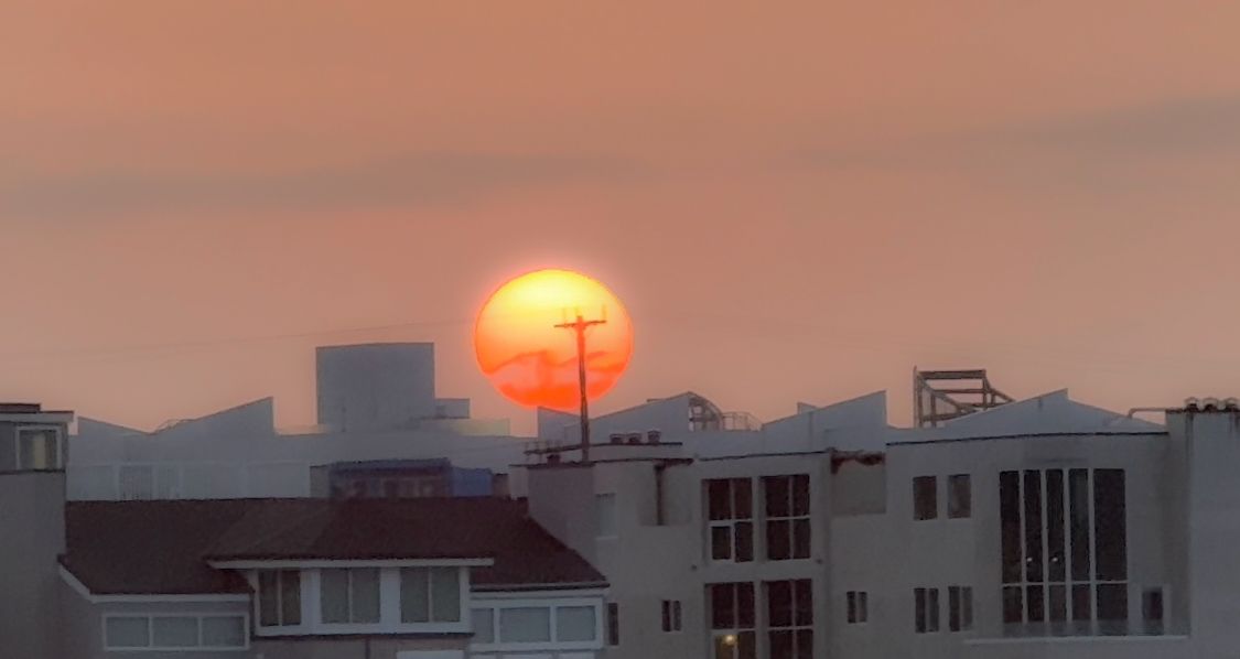 Large orange sun rising behind urban buildings in dusky sky.