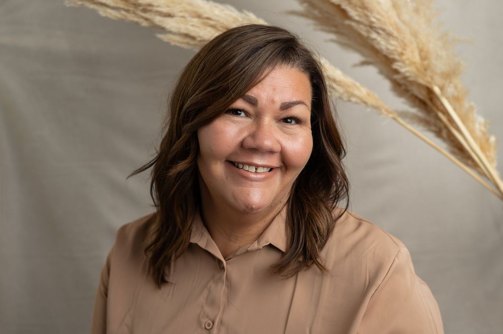Professional portrait with warm lighting featuring brown shirt and decorative pampas grass against neutral backdrop.